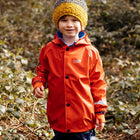 A boy wearing a red Rainy Day Waterproof Jacket standing in a wooded area smiling at the camera. He also has on a yellow and blue bobble hat.